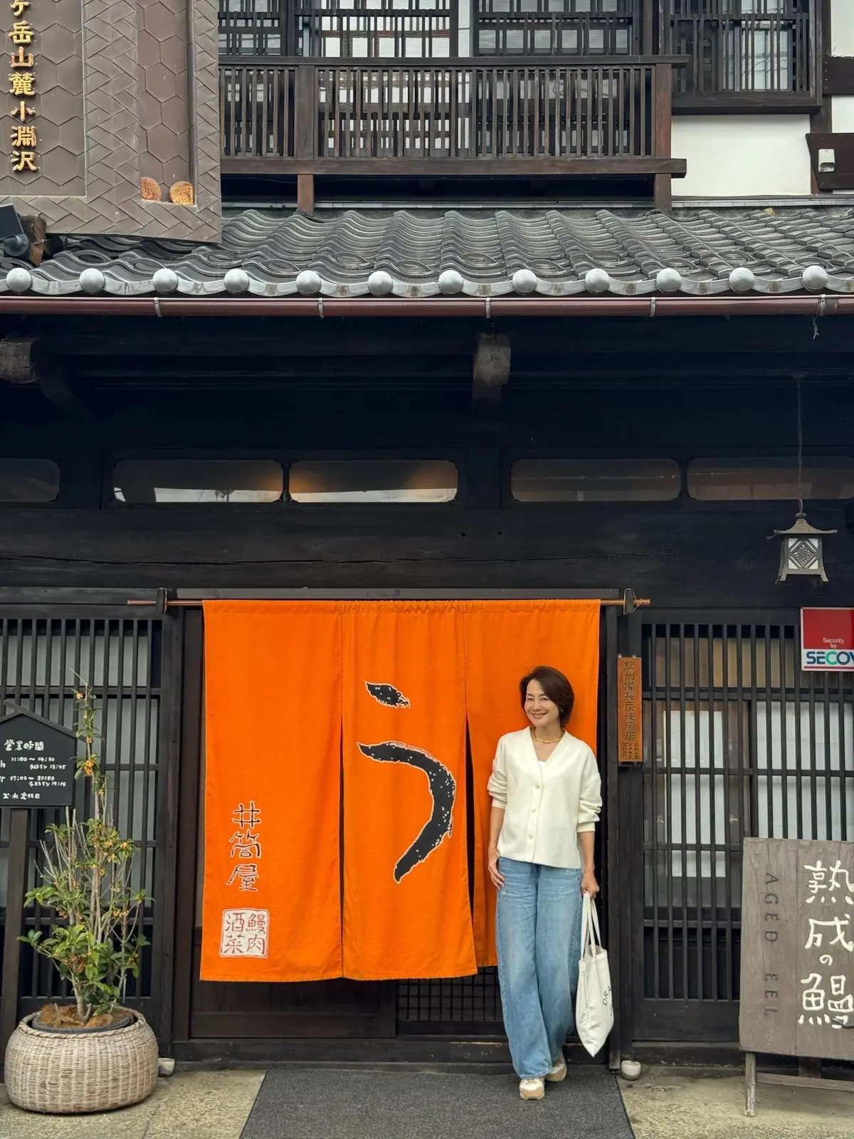 伝統的な木造建築の前に、笑顔の女性2人が立っている。オレンジ色ののれんには黒い文字が描かれ、背景には日本の古い街並みが広がる。