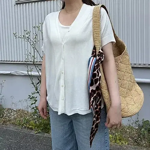 白いボタン付きシャツに、アニマル柄のスカーフを巻いた女性。ベージュの編みバッグを肩にかけ、デニムジーンズを着用。背景にはグレーの建物と緑の植物が見える。