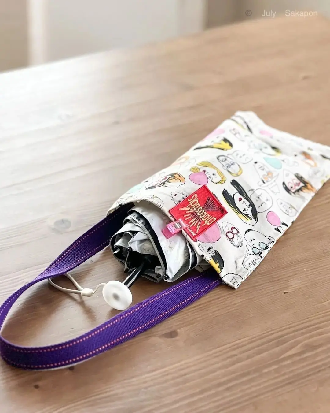 木製のテーブルに置かれた、Chicsticksの傘を収納する白い布製のケース。ケースにはパステルカラーのイラストが描かれ、紫色の持ち手が付いている。傘の柄の一部がケースから覗いている。