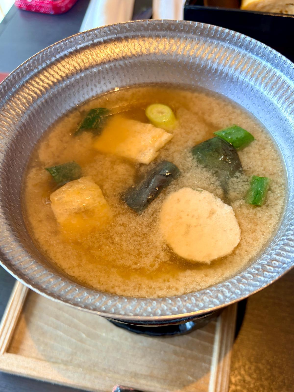 銀色の鍋に入った、薄い茶色のスープ。湯豆腐、ネギ、キノコ、海苔が浮かんでいる。木製のトレイに盛り付けられている。