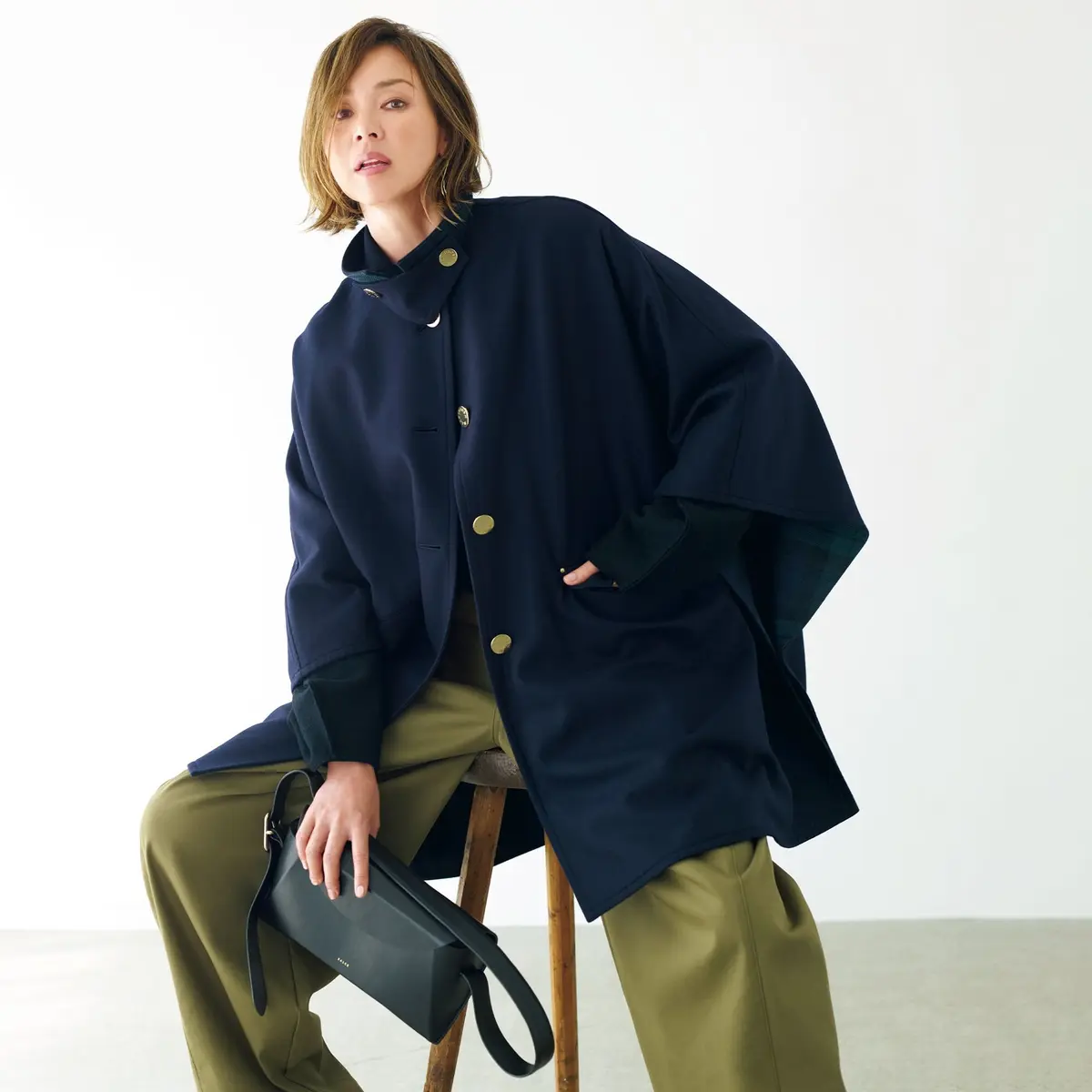 紺色のポンチョコートに、カーキ色のワイドパンツを合わせた女性。右手に黒いバッグを持ち、木製の椅子に寄りかかっている。背景は白。