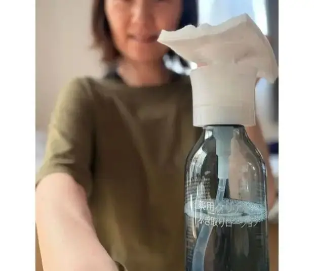 女性が、緑色のTシャツを着て、透明なボトルから取り出す白いシート状の化粧品を見ている。