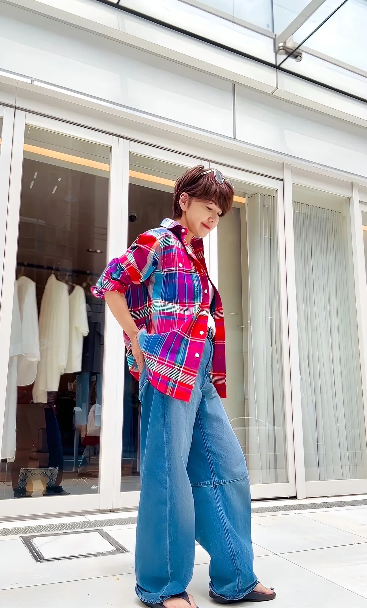 多色チェック柄シャツとデニムパンツを着た女性が、ショッピングモールのガラス窓の前でポーズしています。