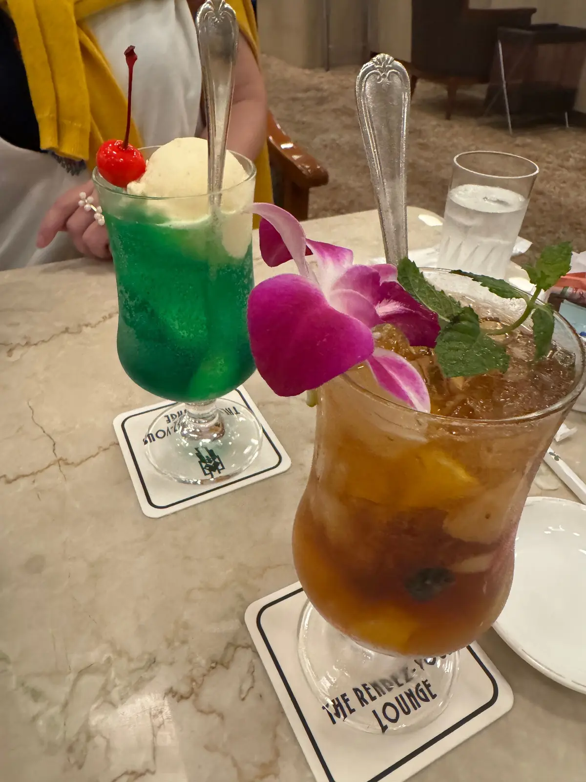 テーブルに置かれた2つのカクテル。左は緑色の液体で丸いチェリーが飾られ、右は黄色がかった液体でピンク色の花が飾られている。両方とも白いコースターの上に置かれ、「The Renazz-Yo Lounge」のロゴが入っている。