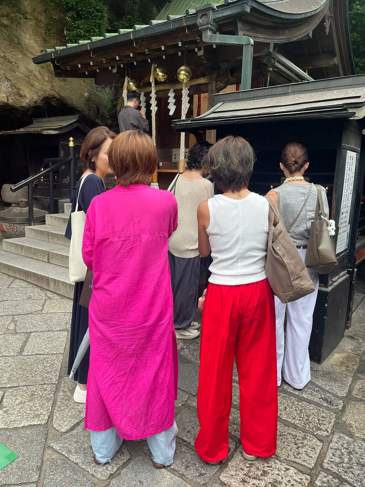 石段に並ぶ５人の観光客が、伝統的な神社建築の入口に集まっている。手荷物を持った女性や、背を向けて祈る男性もいる。銭洗弁天