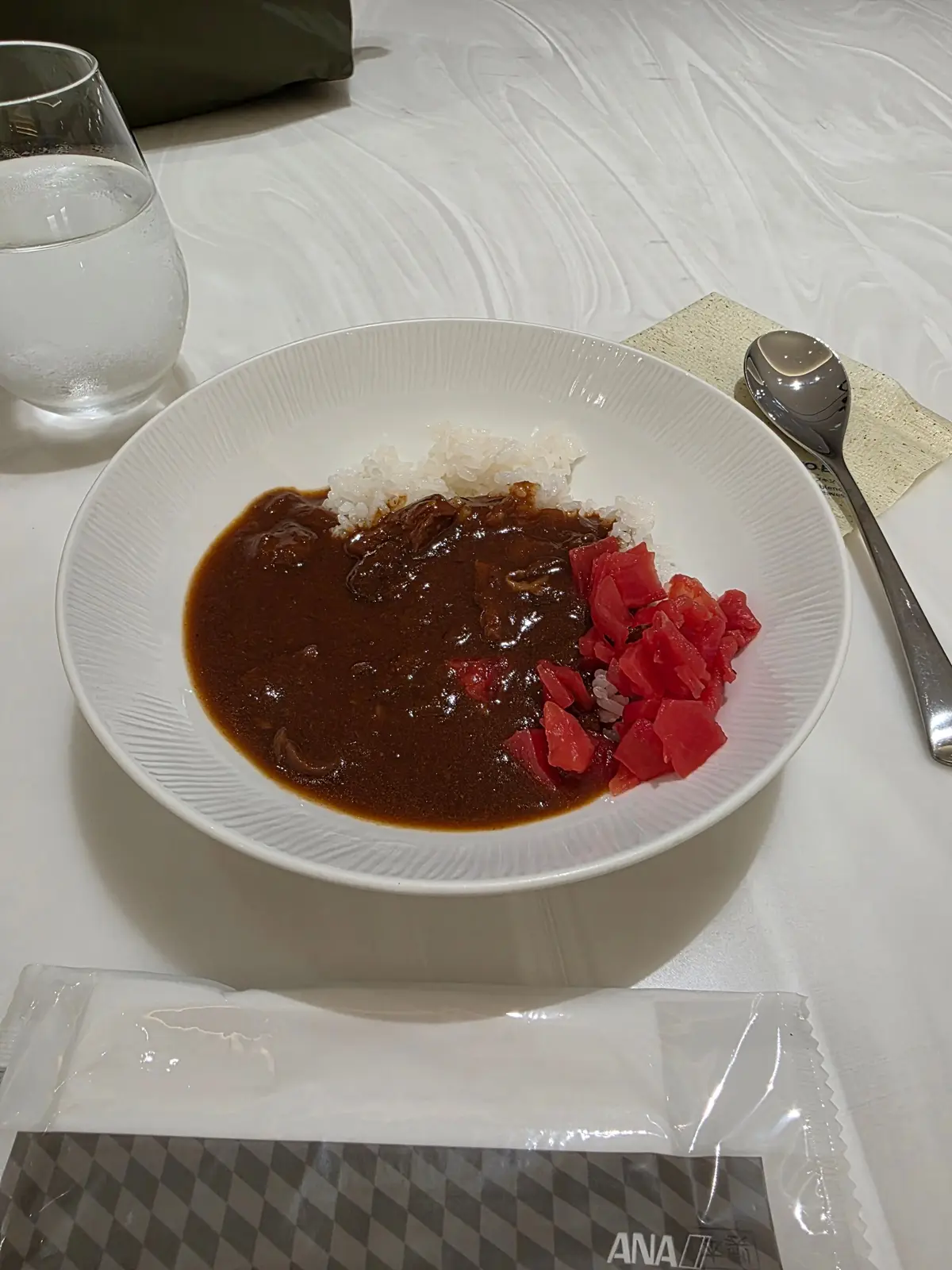 白いボウルに入った日本のカレーライス。ご飯と、赤いダイスカットのトマトが添えられている。ANAのロゴが入ったナプキンが置かれている。