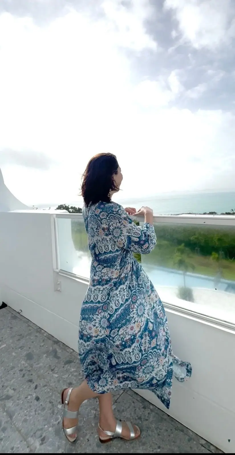 白い手すりにつかまり、海を眺める女性。青と白の柄のロングワンピースを着ており、銀色のサンダルを履いている。背景には緑の植生と、曇り空の海が広がる。