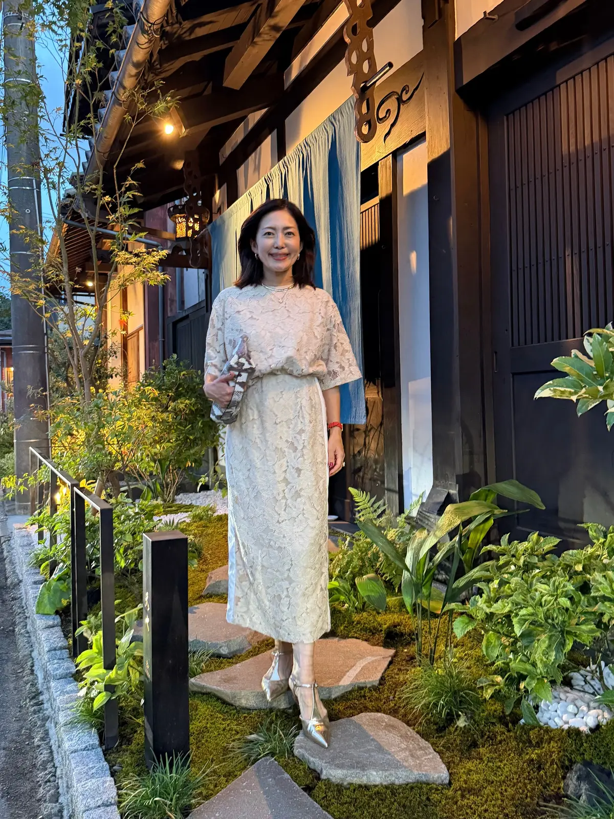上品なレースのワンピースを着た女性が、和風建築の背景に立って微笑んでいる。右手に小ぶりのハンドバッグを持ち、シルバーの靴を履いている。苔むした石庭と植栽が周囲に配置されている。