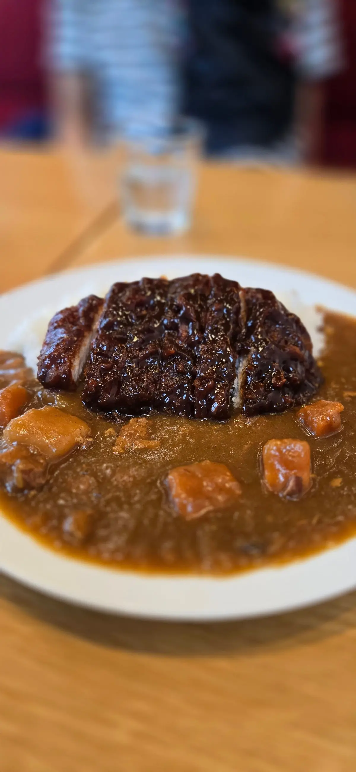 白い皿に、濃いソースをかけた肉と甘い煮物が並んでいる。木製のテーブルに置かれ、背景にはグラスが少しぼやけて見える。