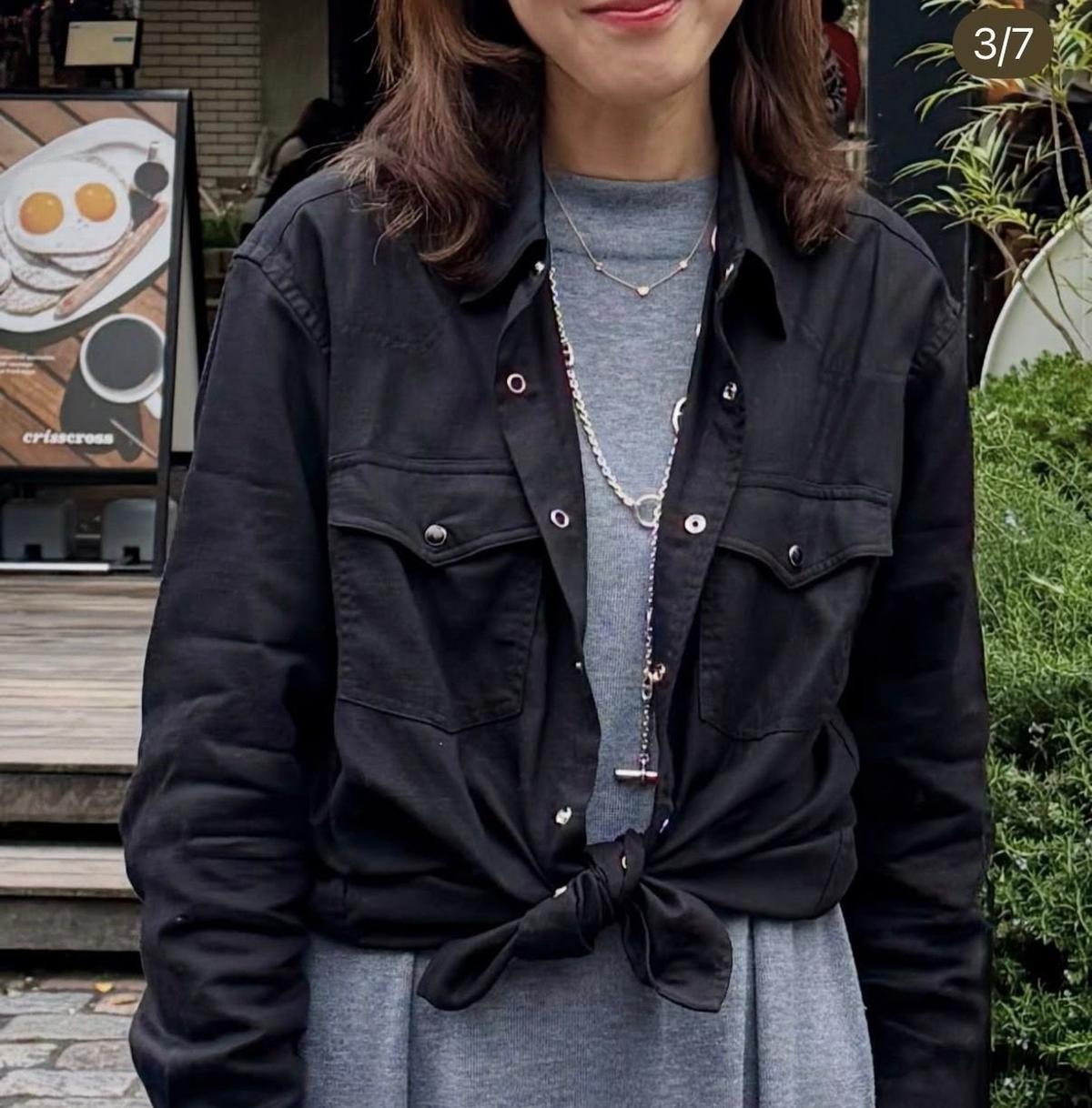 黒いデニムジャケットを羽織った人物。グレーのカットソーの上に着用し、裾を前に結んでいる。シルバーのネックレスを2連で身に着けている。背景にはカフェの看板と緑の植物が見える。