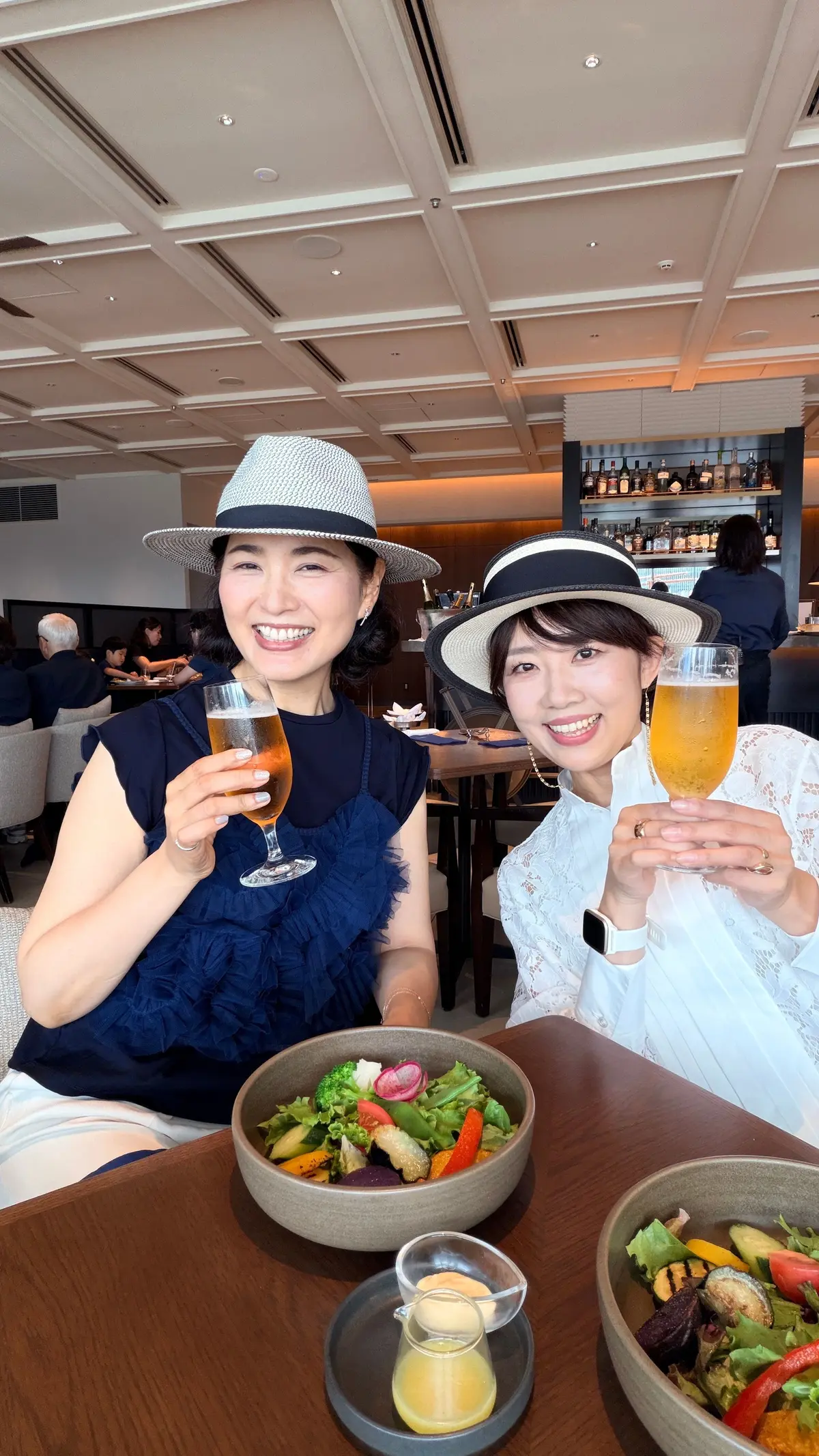 レストランでサラダとビールを手に、笑顔の2人の女性。左は白と黒の帽子、右は黒いリボン付きの帽子をかぶっている。背景にはバーカウンターと客席が見える。