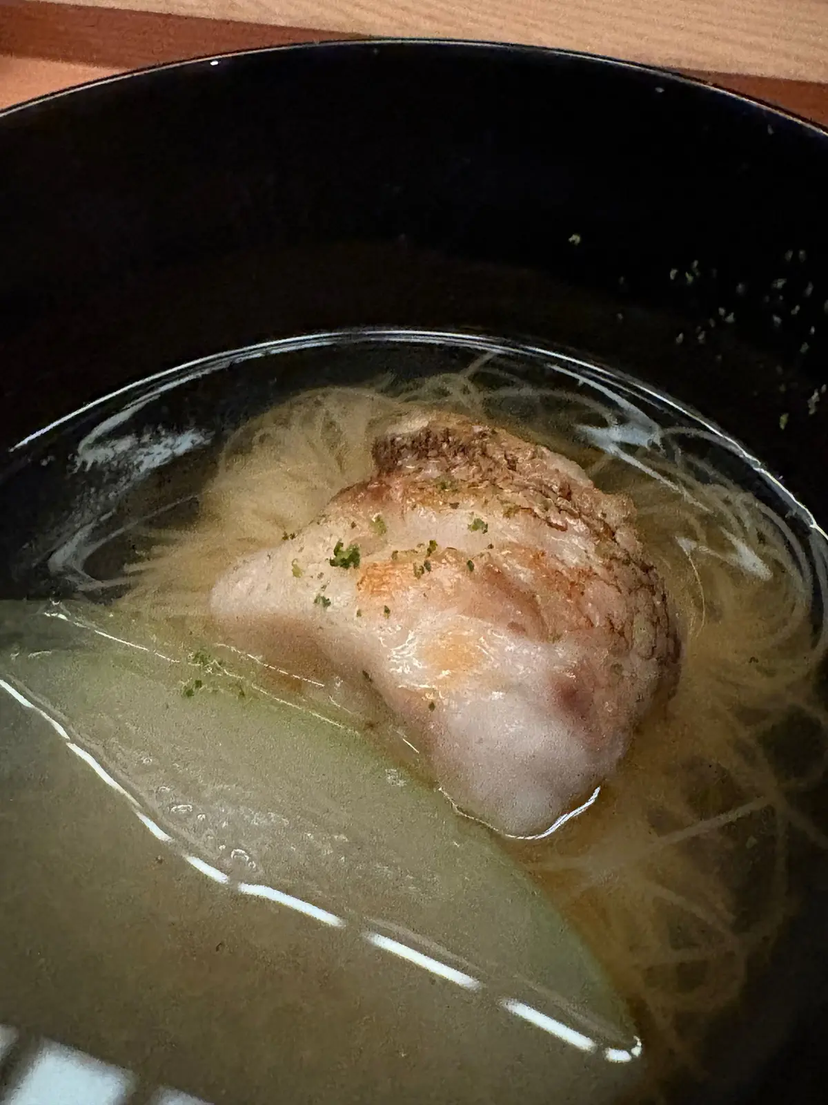 黒い漆器の椀に入った、薄いスープ。スープの中央には、表面に焼き色がついて、白っぽい豚肉が浮かんでいる。スープの表面には油が浮き、細く切られた麺がわずかに見える。