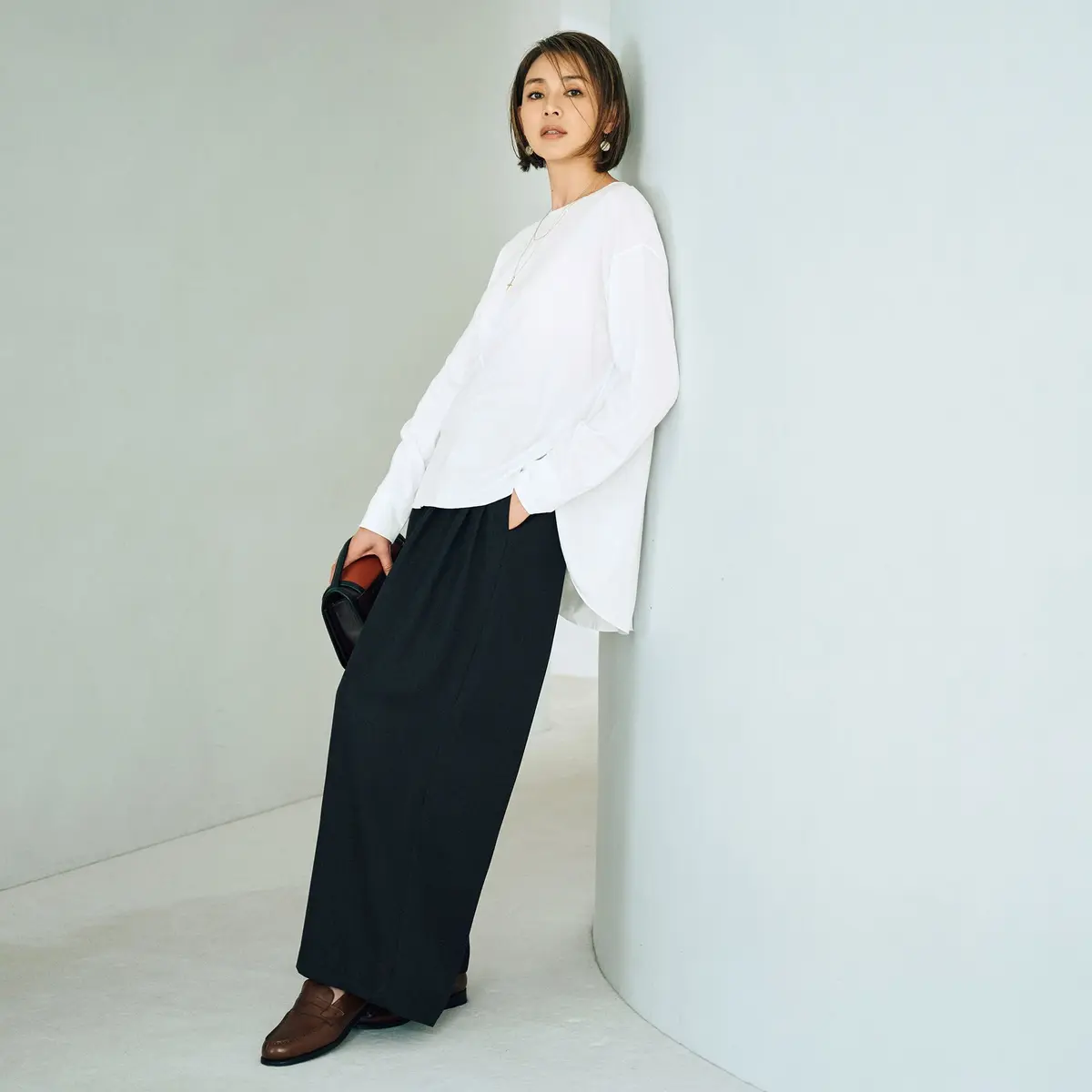 白いシャツに黒いワイドパンツを合わせた女性。茶色のローファーとバッグを身につけ、白い壁に寄りかかっている。