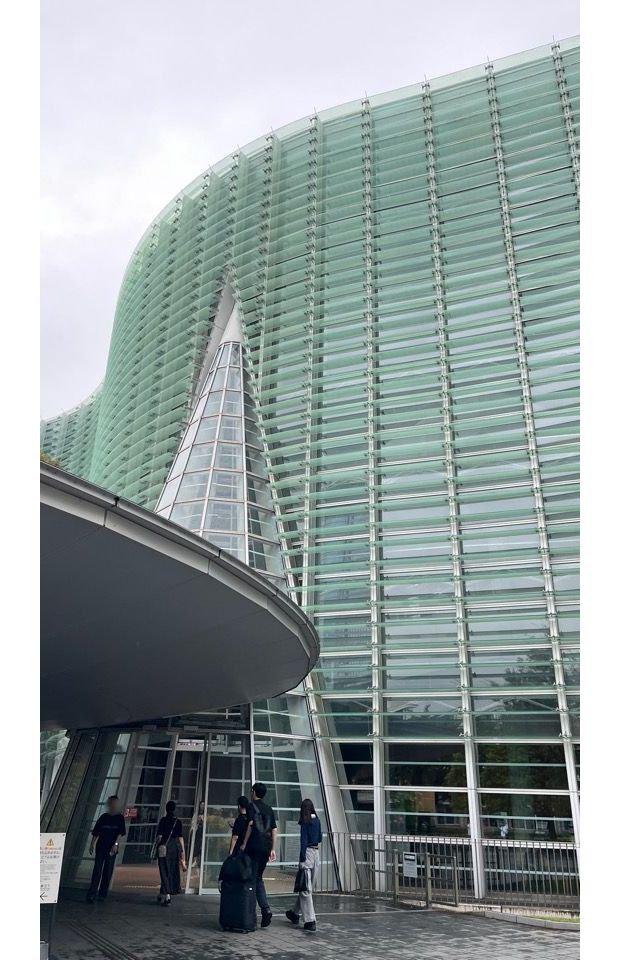 東京の国立新美術館の正門。近代的なガラス張りの建物と、その前に立つ大きな木が特徴的。赤色の案内板が設置され、開館時間が記載されている。