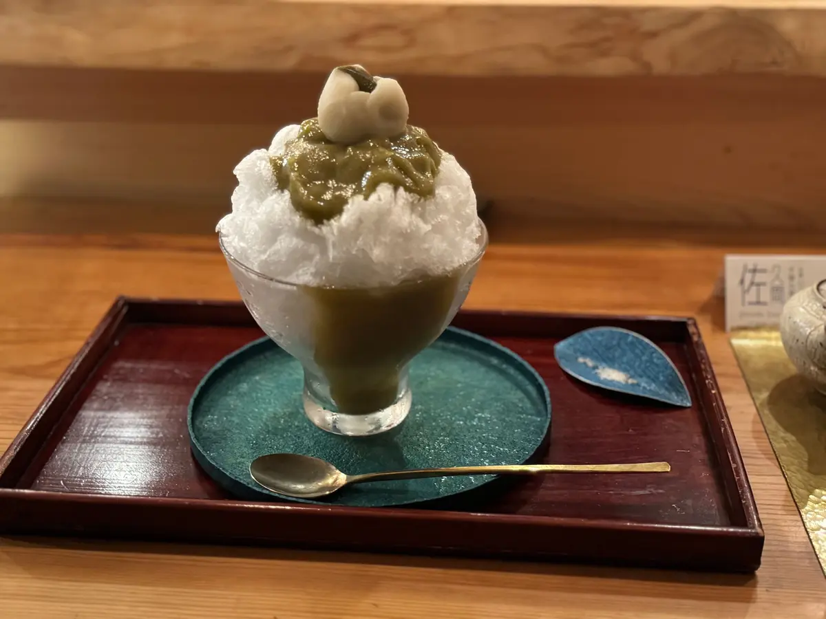 ガラスの器に入ったかき氷に、抹茶色のソースと白玉が乗ったデザート。木製のトレイに置かれ、スプーンが添えられている。