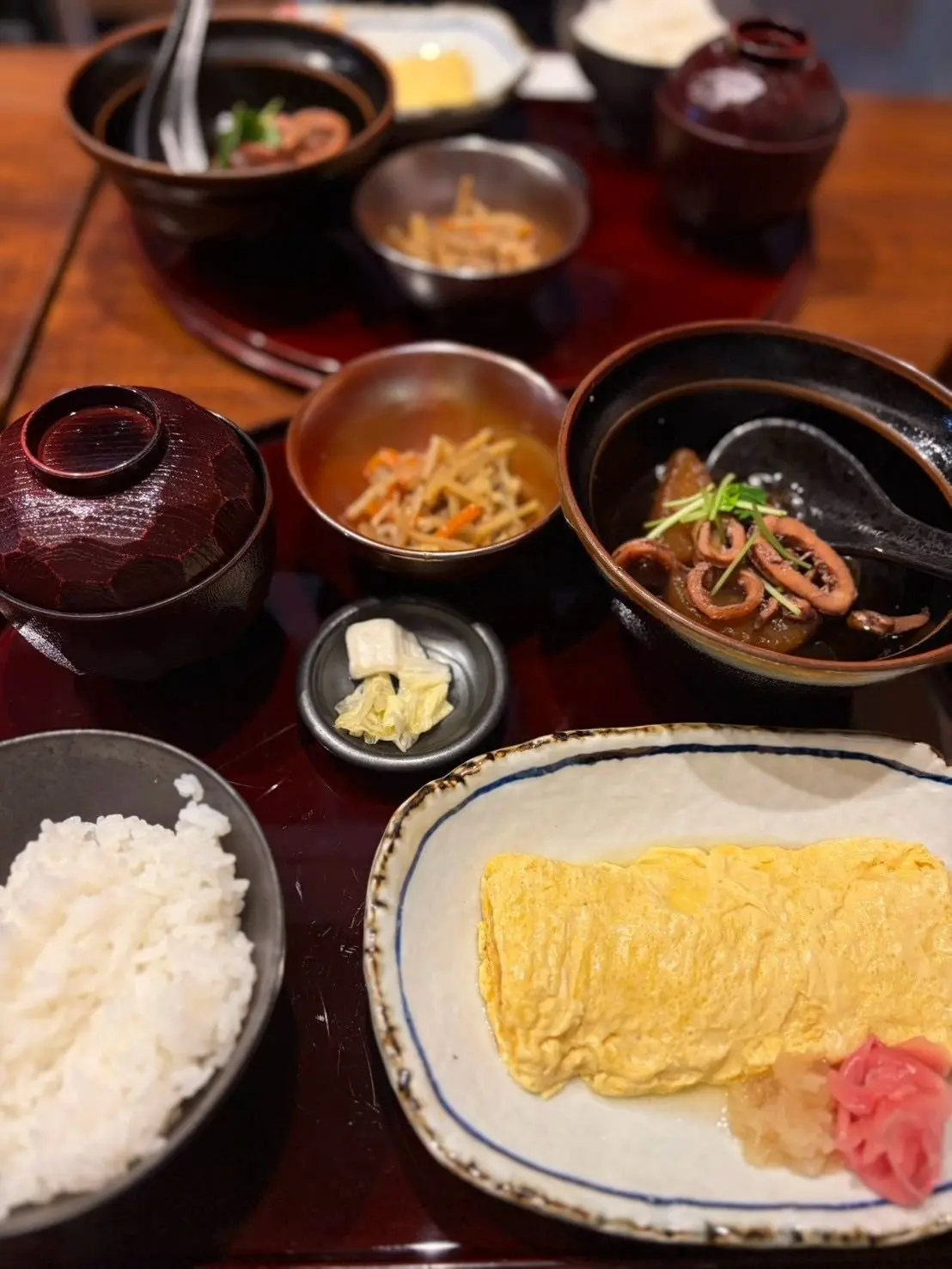 木製のテーブルに盛り付けられた、日本の定食。白米、卵焼き、きんぴらごぼう、豚肉の角煮、香の物など、様々な料理が碗や皿に盛り付けられている。