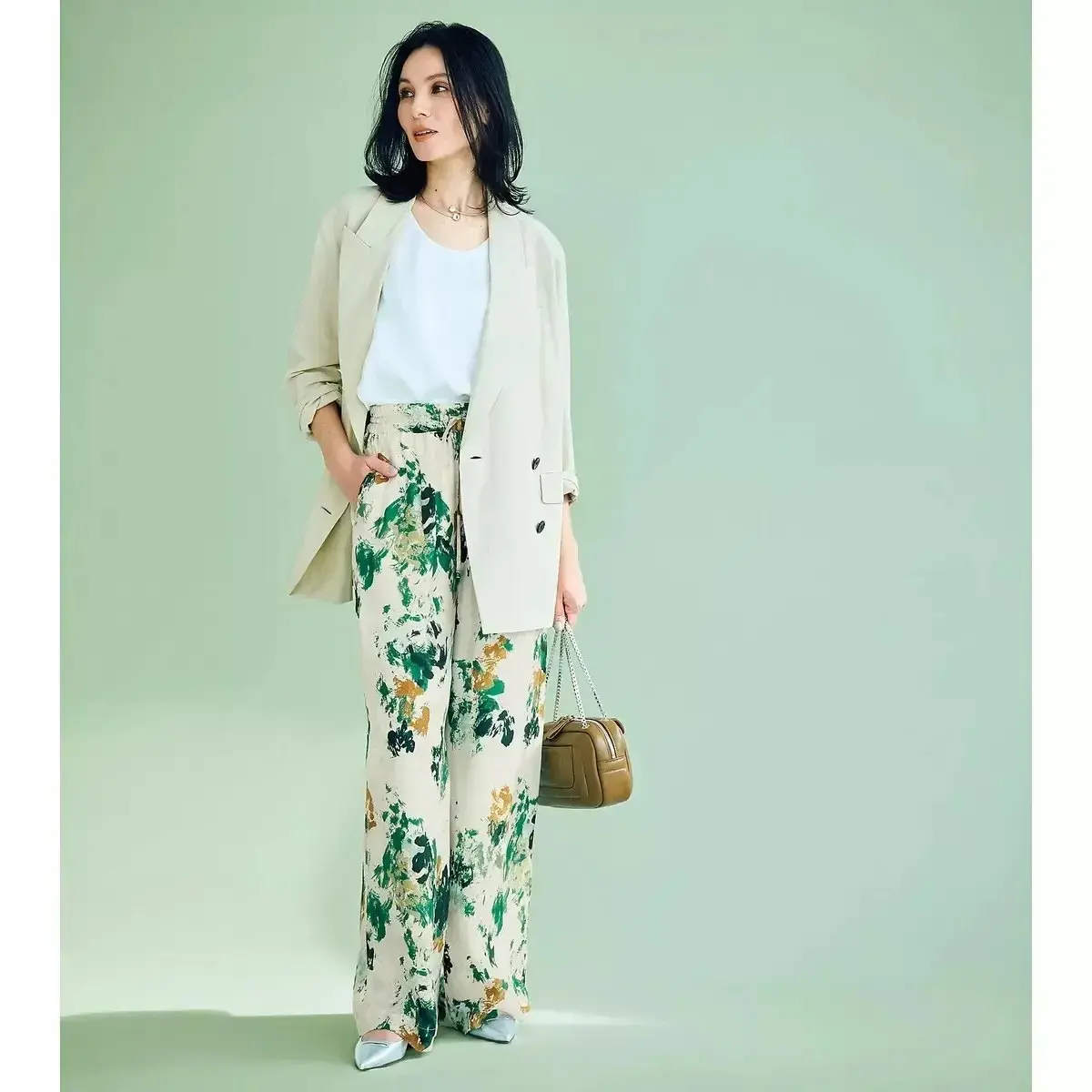 ベージュのブレザージャケットと白のトップスを着た女性が、緑と黄色の花柄のズボンを履き、右手に茶色のショルダーバッグを下げて立っている。