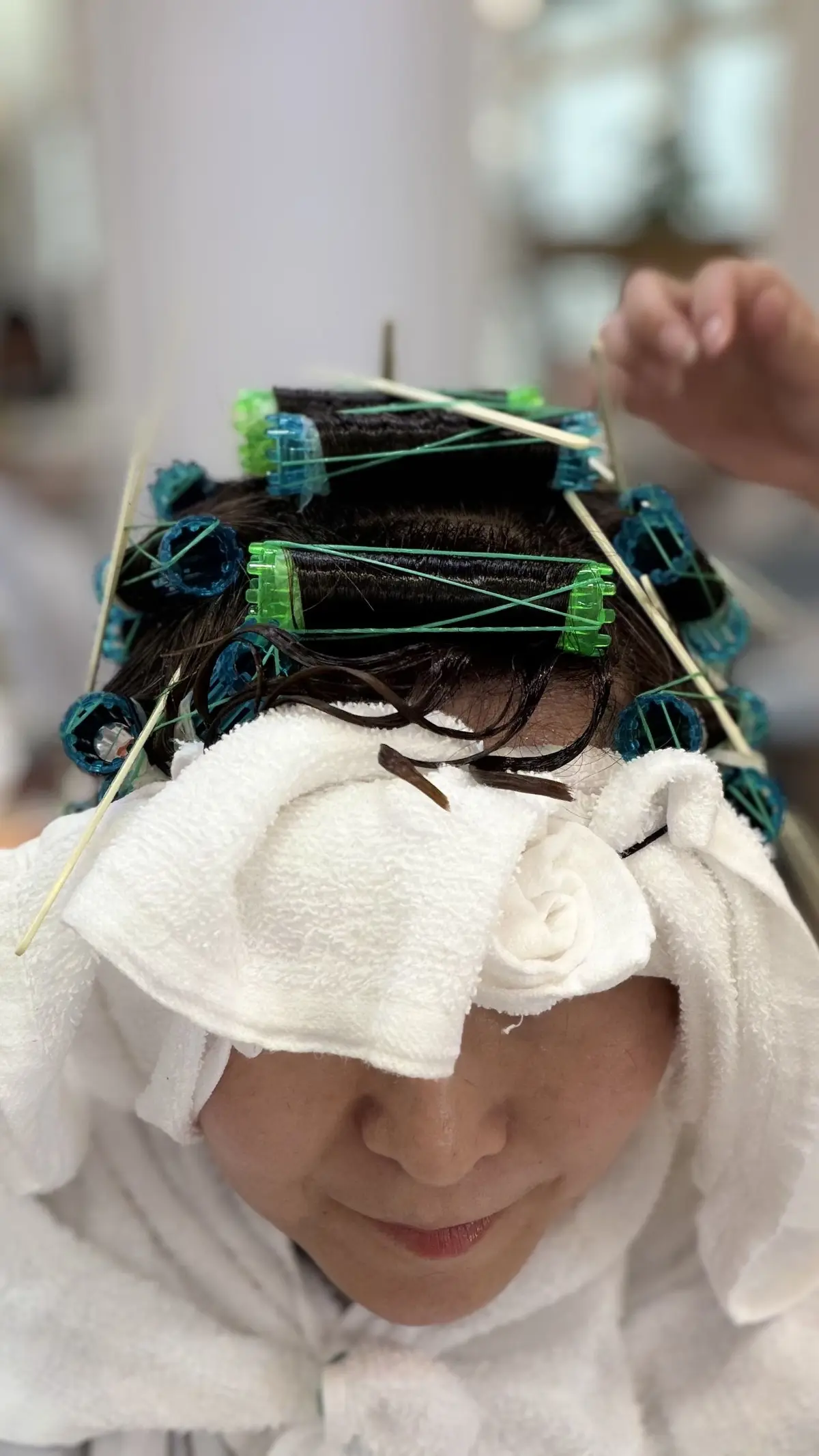 白のタオルを頭に巻き、緑と青のヘアカーラーを数個入れた子供。木製のピンで固定され、顔の一部がタオルで隠れている。