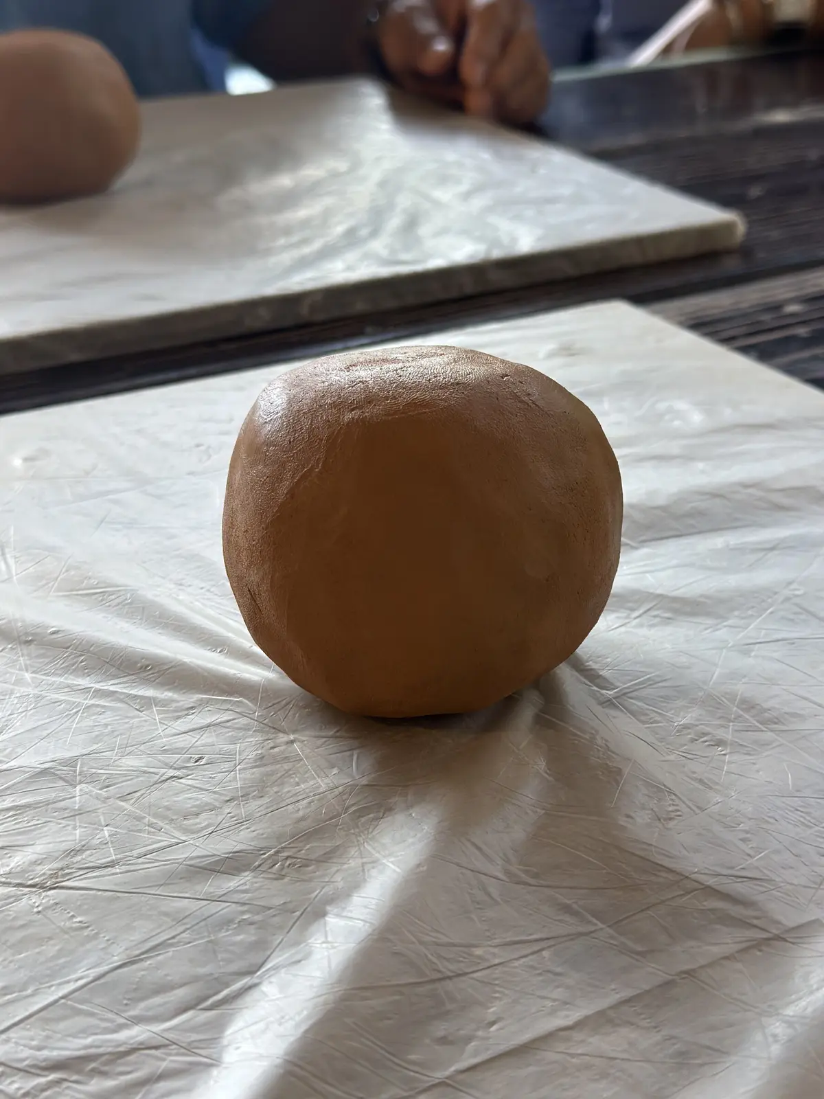 茶色の粘土が丸い塊の形をして、白いシートの上に置かれている。背景には、別の粘土の塊と作業台が見える。