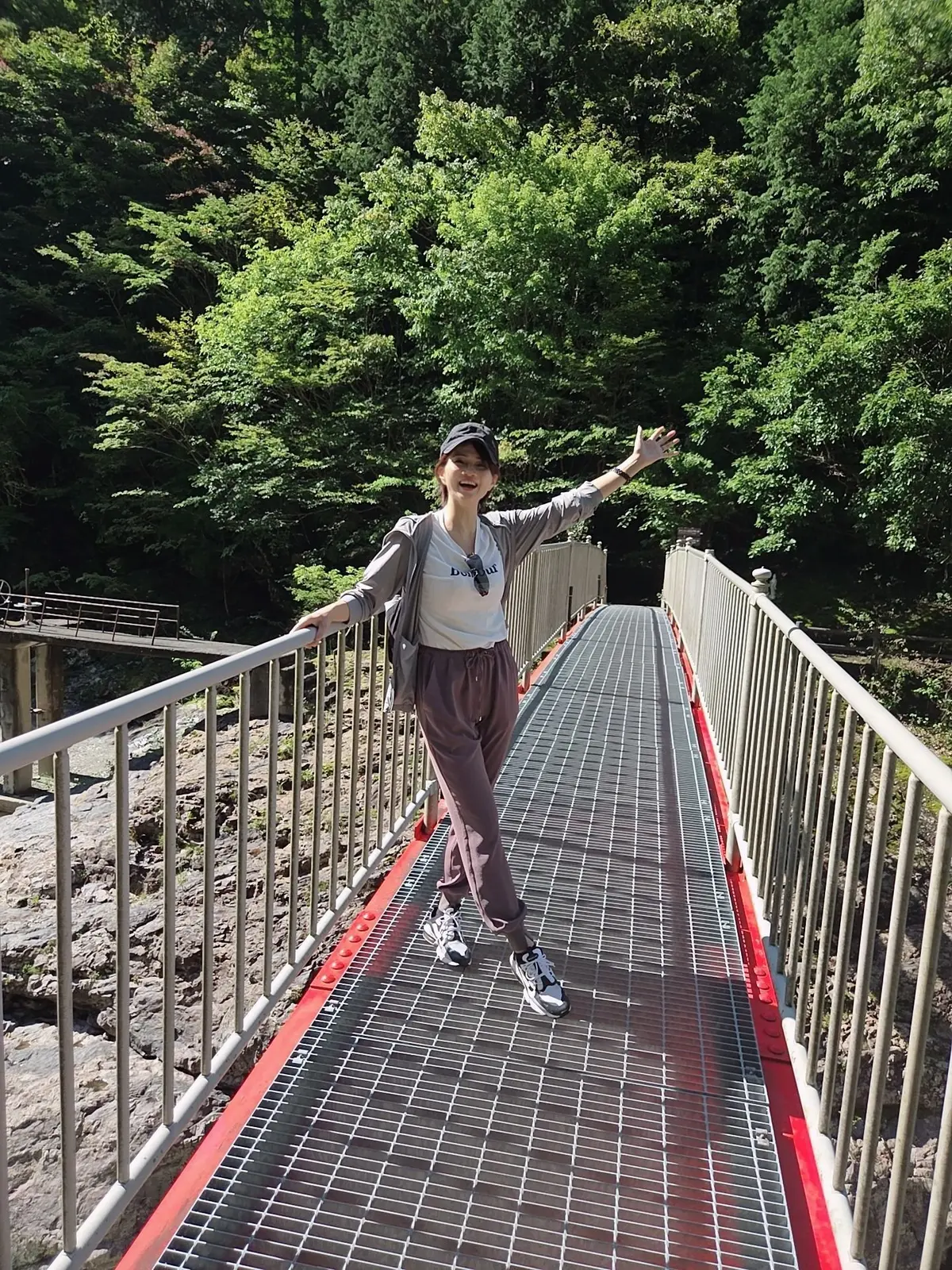 緑豊かな木々を背景に、赤い手すりの橋の上でポーズをとる女性。グレーのトップスにカーディガン、スウェットパンツ、スニーカーを着用。両手を広げ、笑顔を見せている。