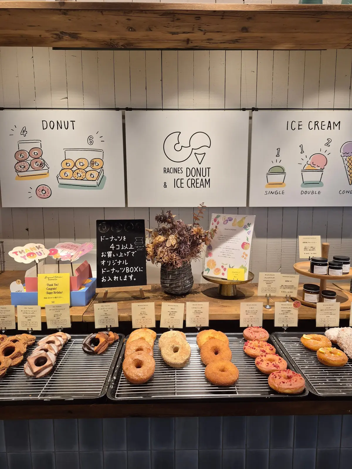 Racines Donut & Ice Creamの店舗の様子。グレーのショーケースに、様々な種類のドーナツが並んでいる。背景には、ドーナツやアイスクリームのメニューが書かれた木製の看板が設置されている。