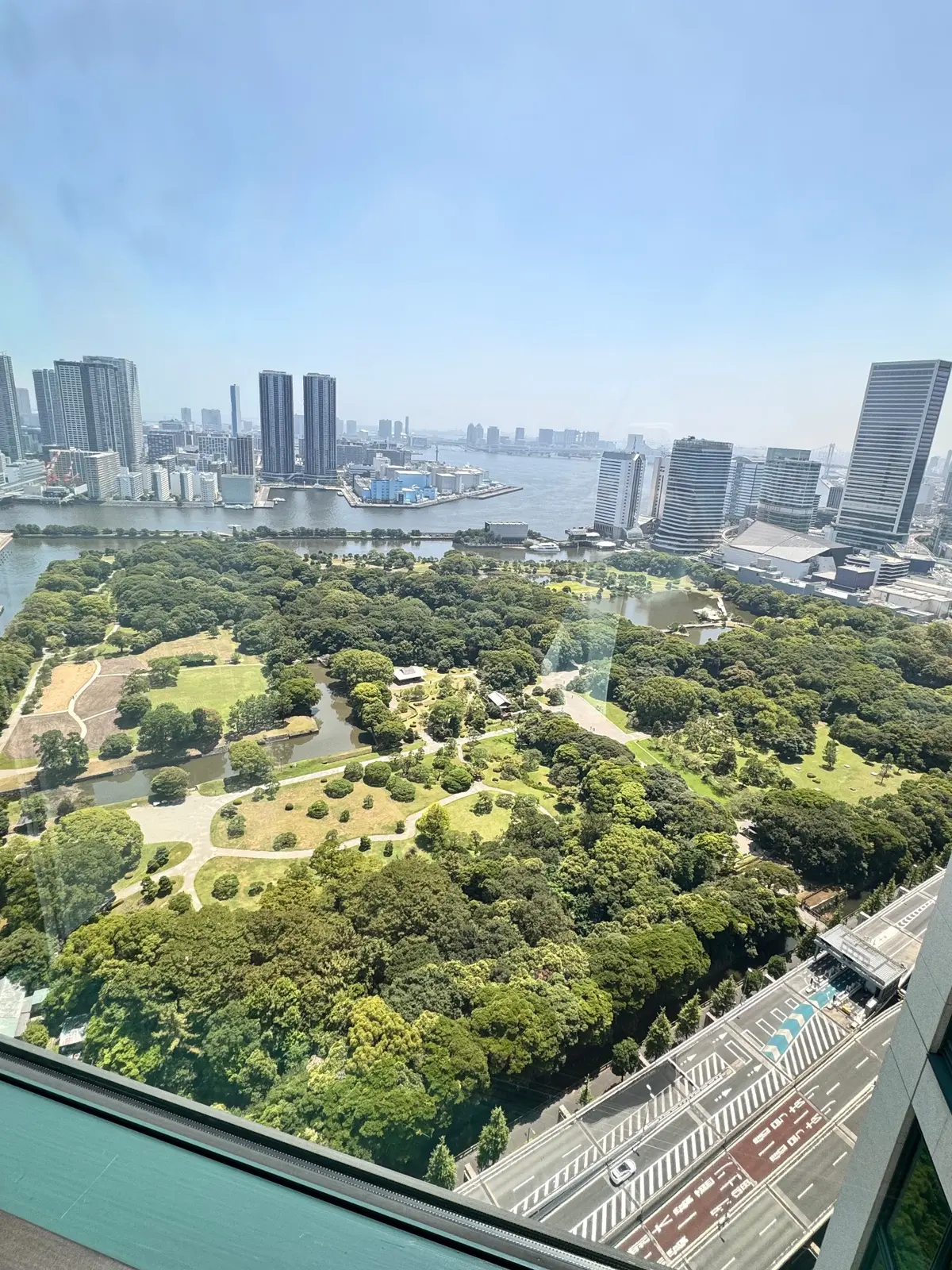 高層ビルからの眺め。緑豊かな公園と、水辺に沿って建ち並ぶ高層ビル群が広がる都市の風景。晴天の青空の下、道路や水面がキラキラと輝いている。