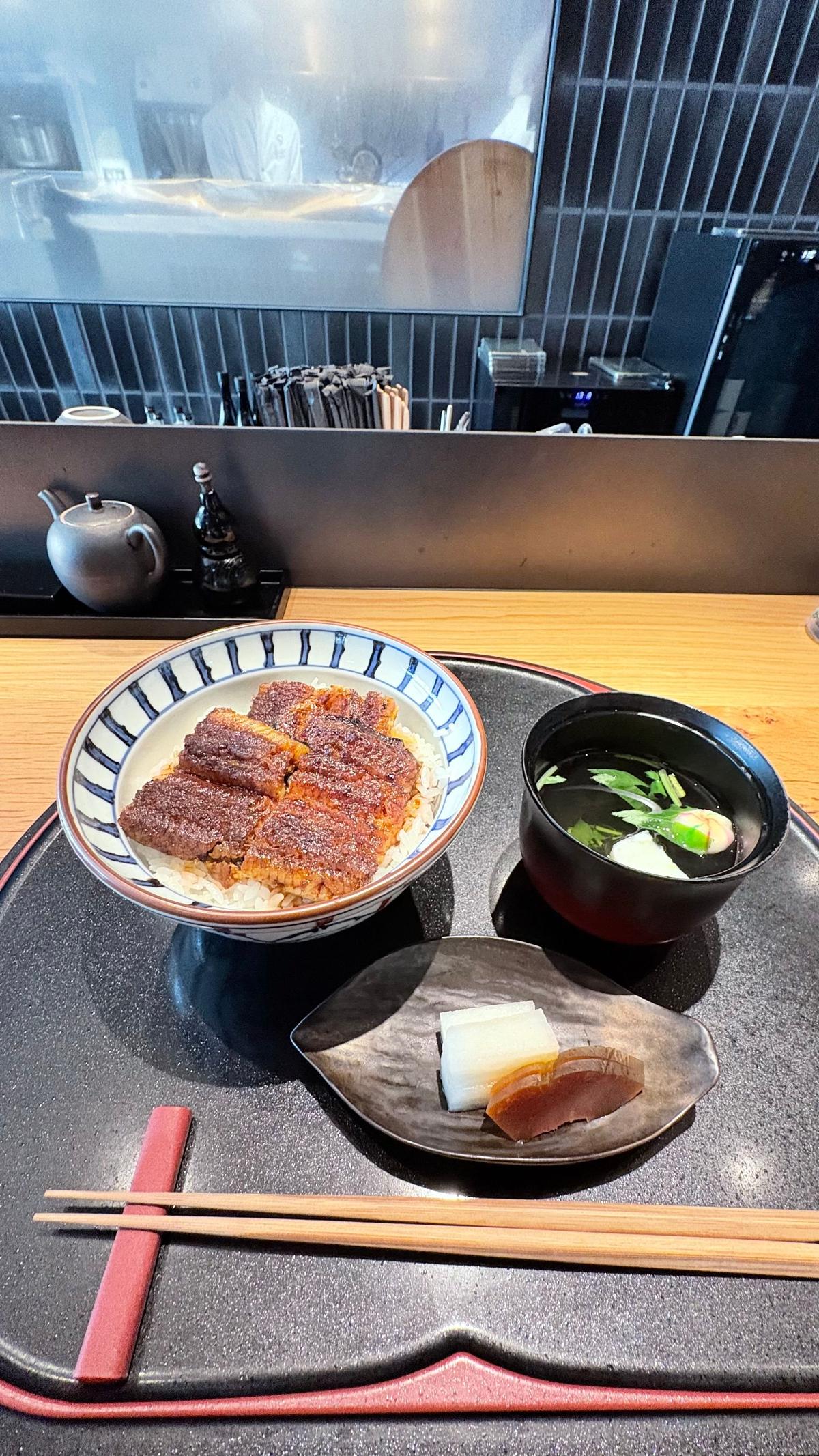 黒いトレイに盛り付けられた、ご飯とカツ丼、味噌汁、漬物のセット。カツ丼は白米の上に揚げた豚肉が乗っており、味噌汁にはわかめとネギが入っている。