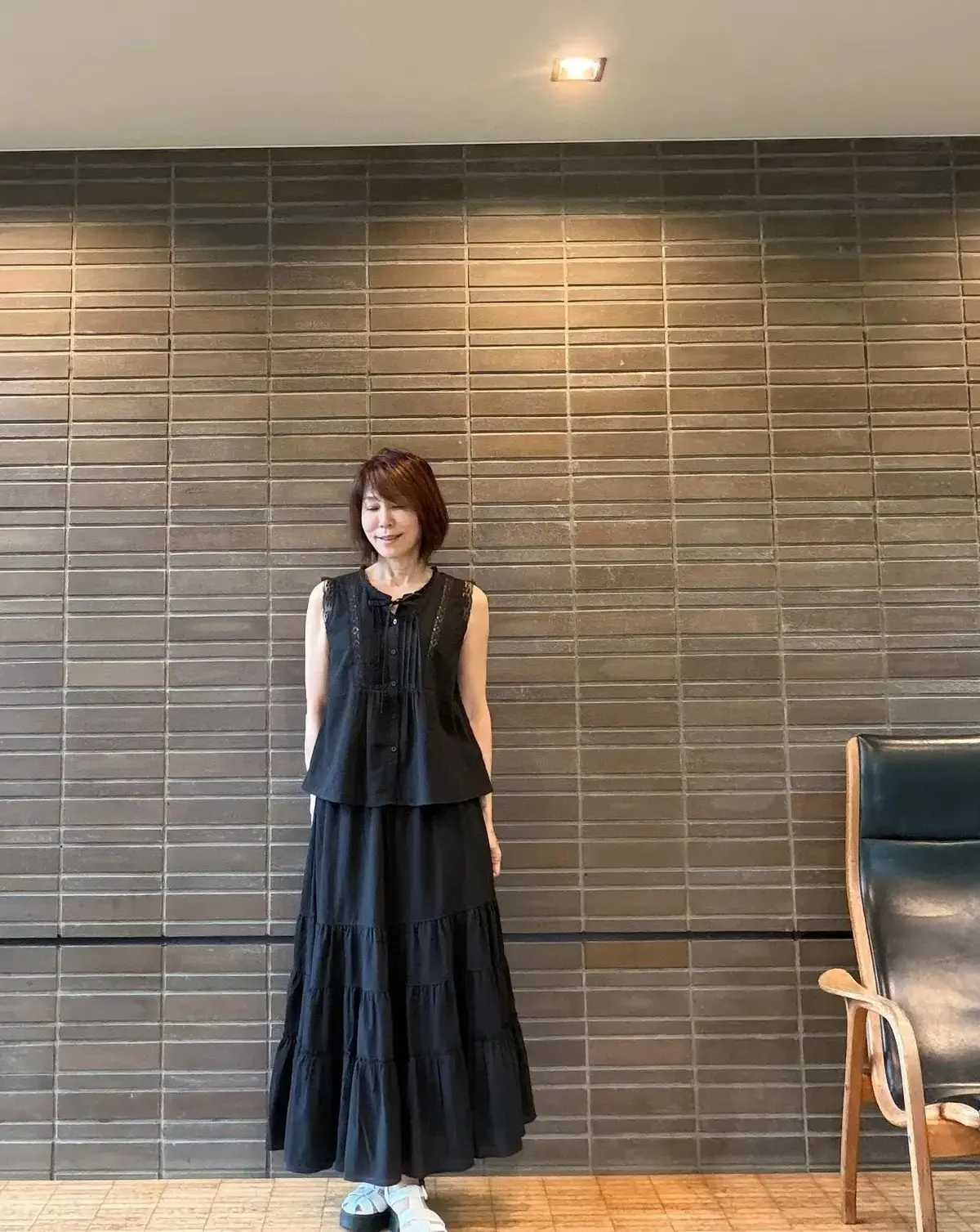 茶色のタイル壁に、黒のフレアスカートとチュニックを着た女性が立っている。右側に木製の椅子がある。