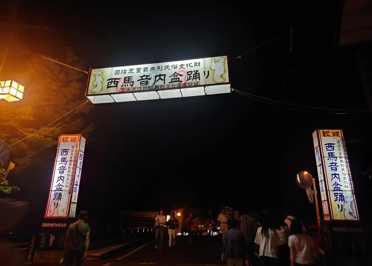 夜の暗闇に、提灯と看板で彩られた「西馬音内金照り」の入り口。黄色と赤の看板が目立ち、提灯が左右に並んでいる。手前に人影が見える。