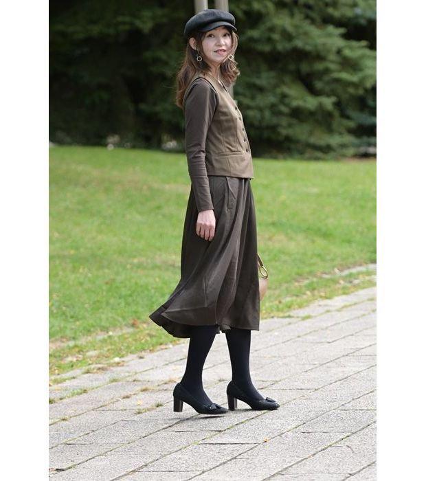 黒の帽子を被った女性が、緑の芝生の横の歩道に立っている。茶色の長袖ワンピースにブラウンのベストと、黒のタイツとパンプスを着用。シンプルなイヤリングをつけ、茶色のハンドバッグを持ってている。