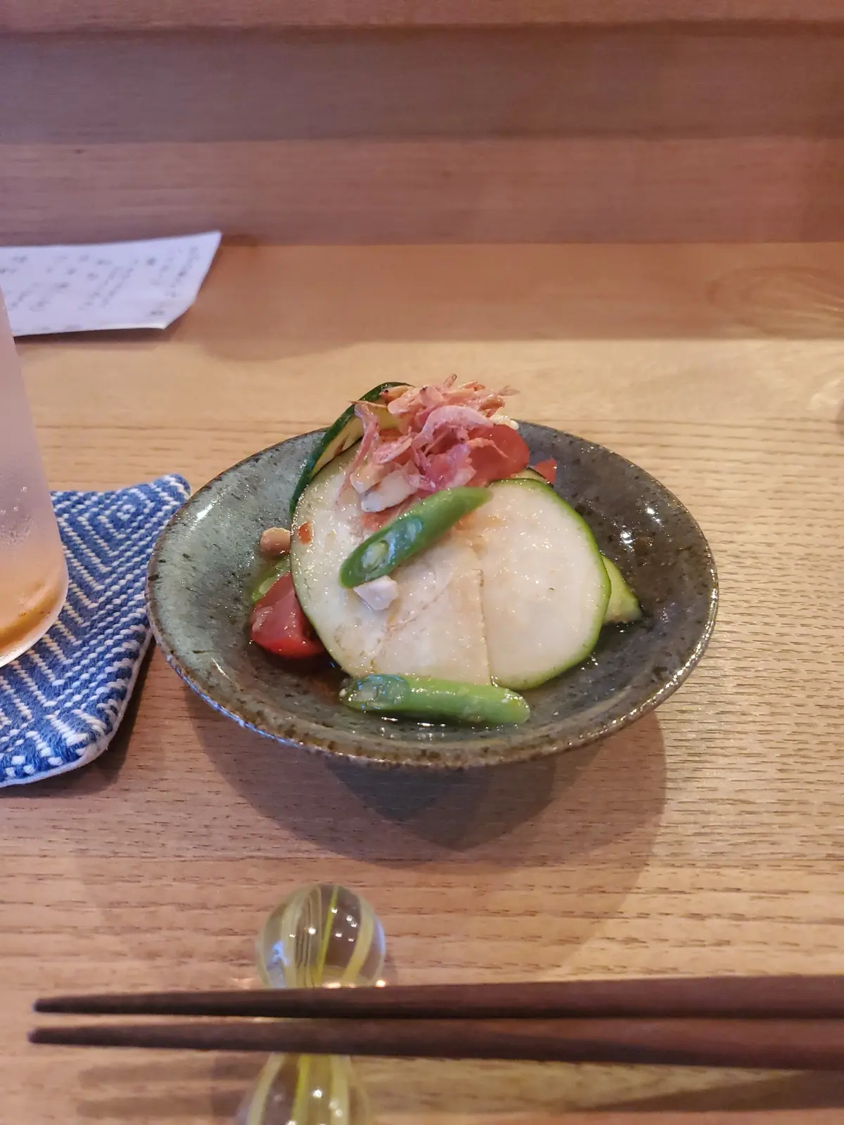 木製のテーブルに置かれた、小皿に入った和食。白身魚の薄切り、キュウリ、トマト、エディブルフラワー、赤いネギなどが彩り豊かに盛り付けられている。青と白のランチョンマットと、木製の箸が添えられている。