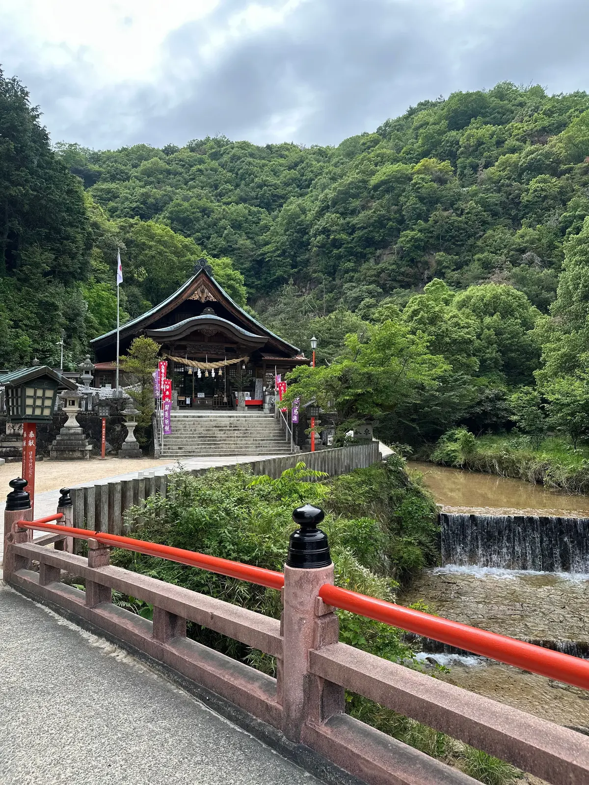 緑豊かな山に囲まれた川沿いの神社で、赤い手すりがあり、石段を登った場所に伝統的な建築の社殿が立つ