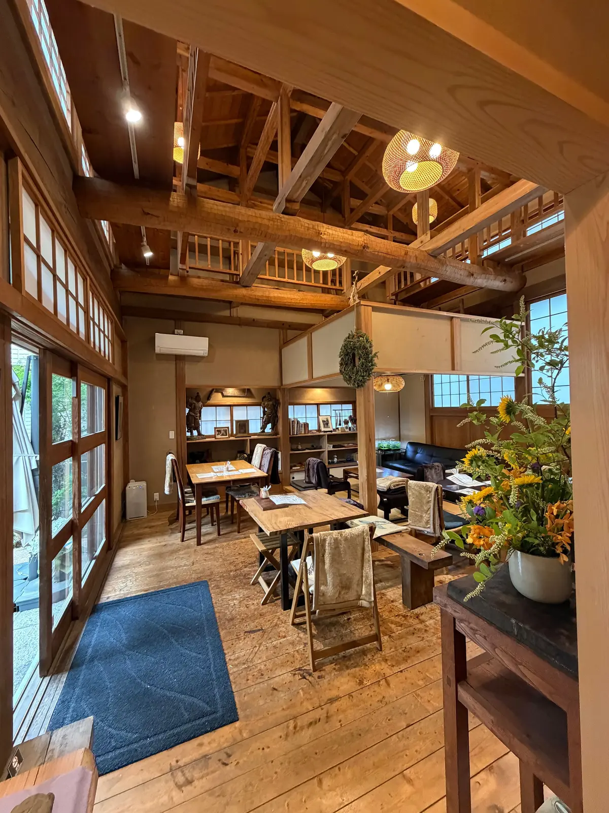 木造の広いリビングルームで、木製の天井と大窓から自然光が差し込む。木製のダイニングテーブルと椅子、ソファ、花の入った花瓶があり、青い敷物が床に敷かれている。