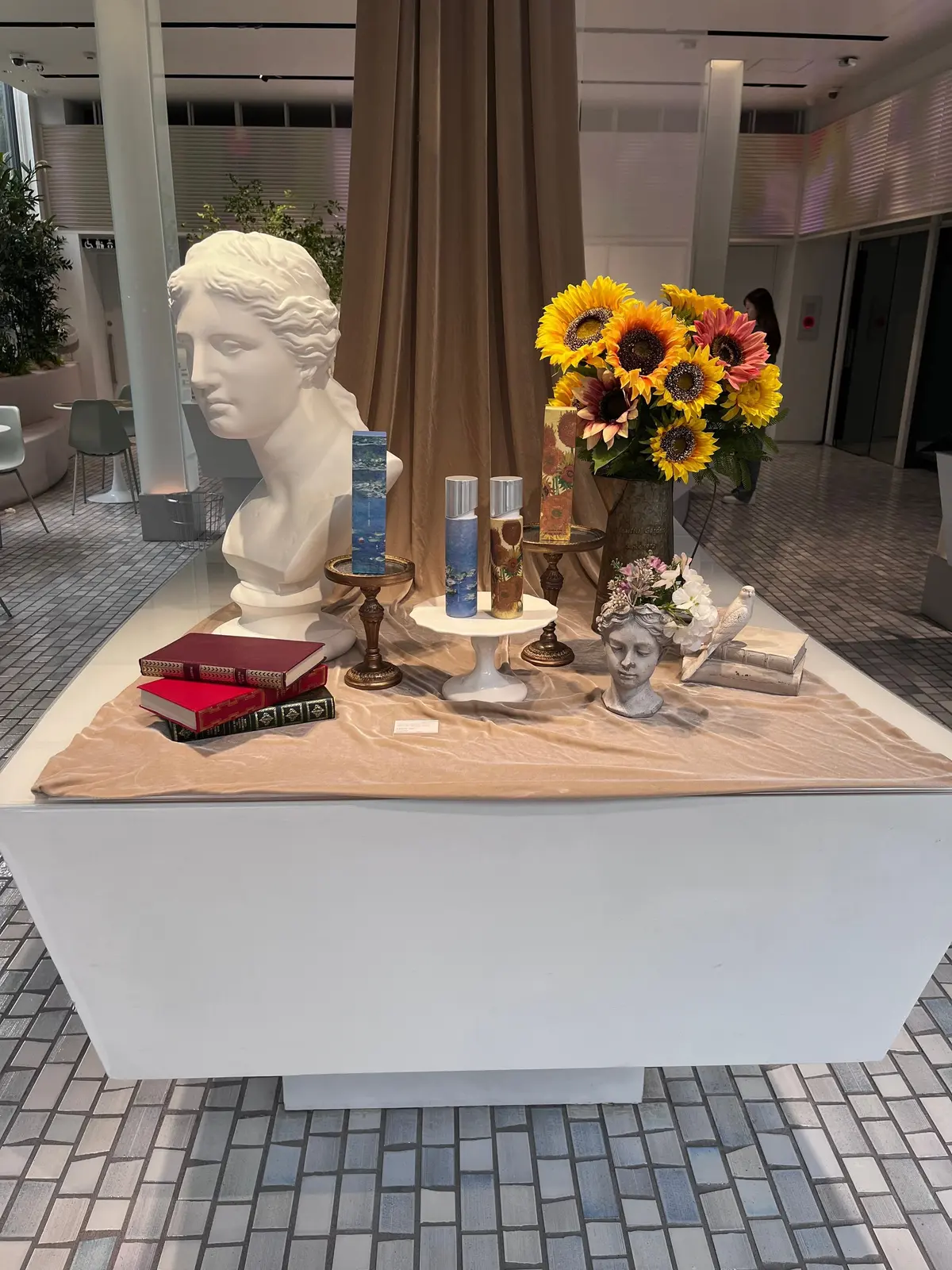 白いテーブルの上に、彫像、花束、本、装飾品が配置されたディスプレイ。左には大理石の胸像、右にはヒマワリの花束が置かれ、中央には本やキャンドルなどが並んでいる。背景にはベージュのカーテンがかかっている。