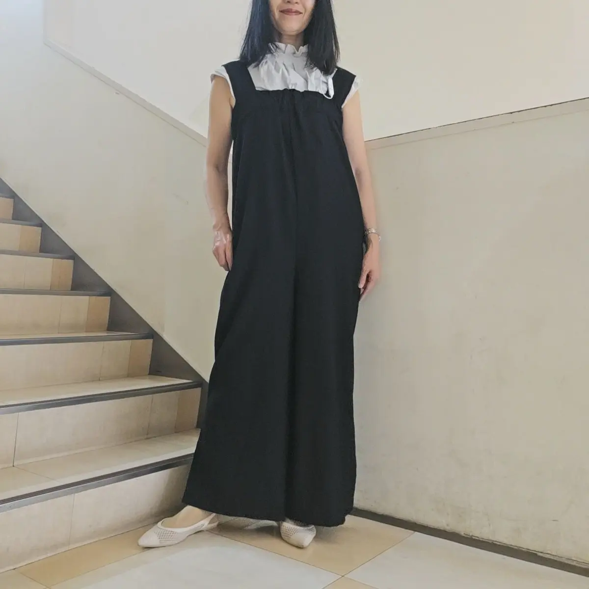黒いオーバーオール型のロングワンピースを着た女性。白いフリルシャツを中に着用し、白いメリージェーンシューズを履いている。背景には階段がある。