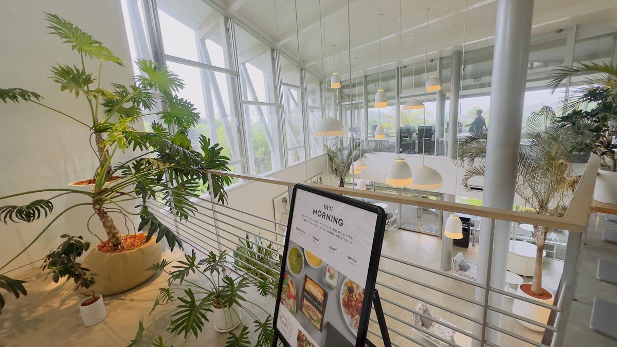 白を基調とした明るい空間に、観葉植物が置かれたカフェの様子。窓から外の景色が見え、吹き抜けの空間にはペンダントライトが吊り下げられている。手前にメニューボードが置かれている。