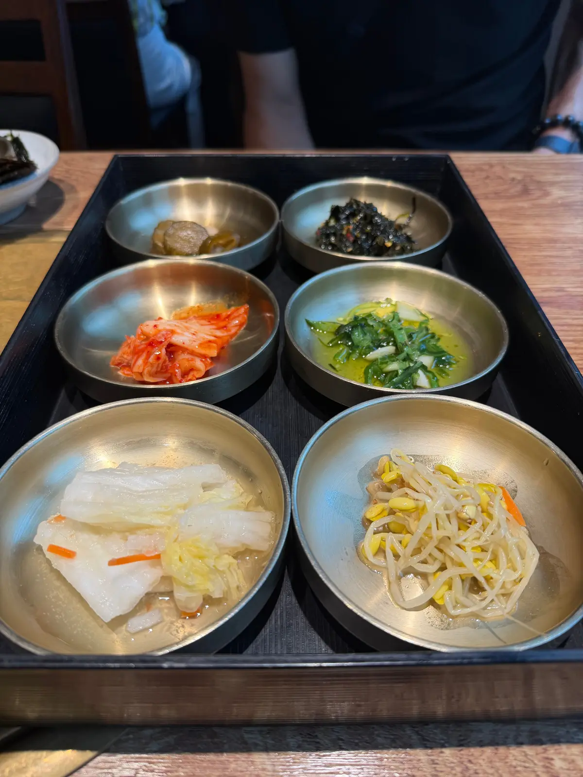木製トレイに並んだ六つの金属ボウルに、海藻、キムチ、ナムル、刺身、麺などの韓国の前菜が盛られている