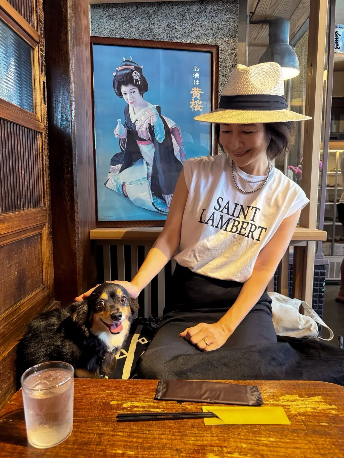 レストランのテーブル席で、若い女性が茶色の帽子をかぶり、白いTシャツを着て犬を撫でている。背景には日本の女性の絵画が飾られている。テーブルにはグラスとマスクが置かれている。