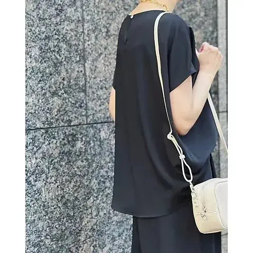 黒いトップスとパンツを着用した女性が、白いカメラバッグを肩にかけ立っている。背景はグレーの石材模様の壁。