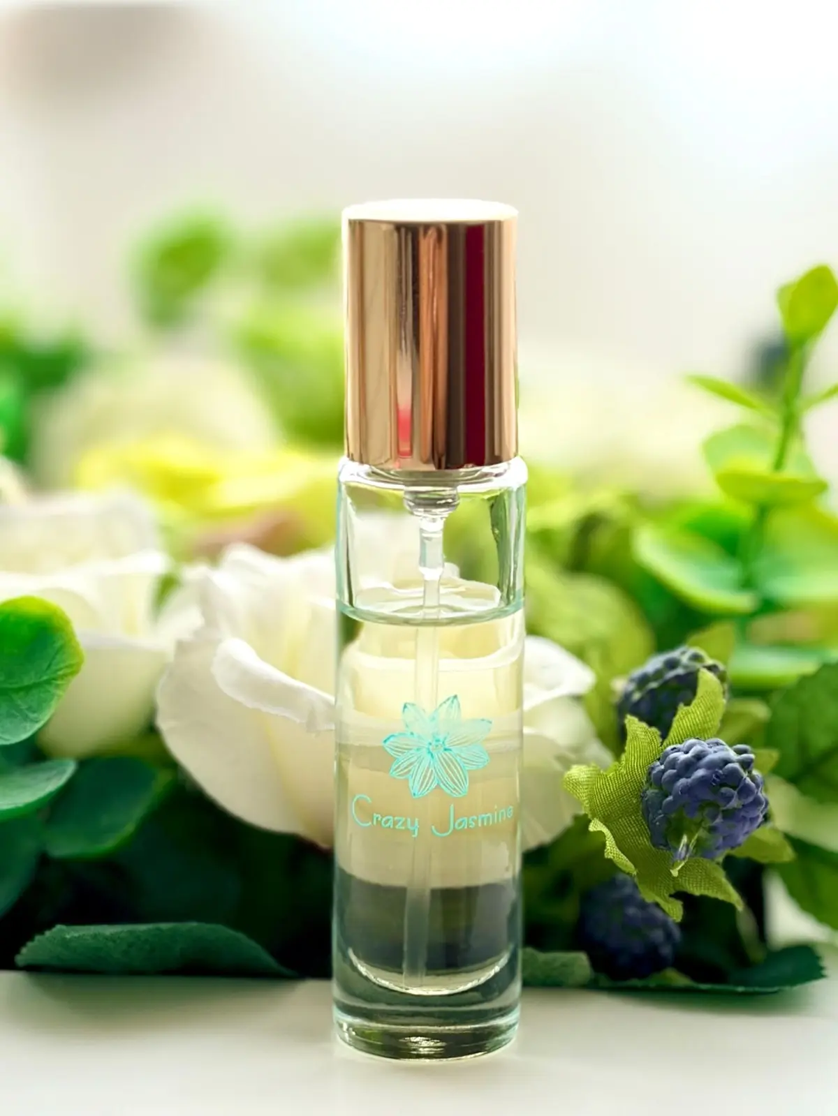 ガラス製の香水ボトルで、銅色のキャップと青い花のデザインが特徴。ボトルには「Crazy Jasmine」という文字が書かれている。背景には白いバラと青い花が配置されている。