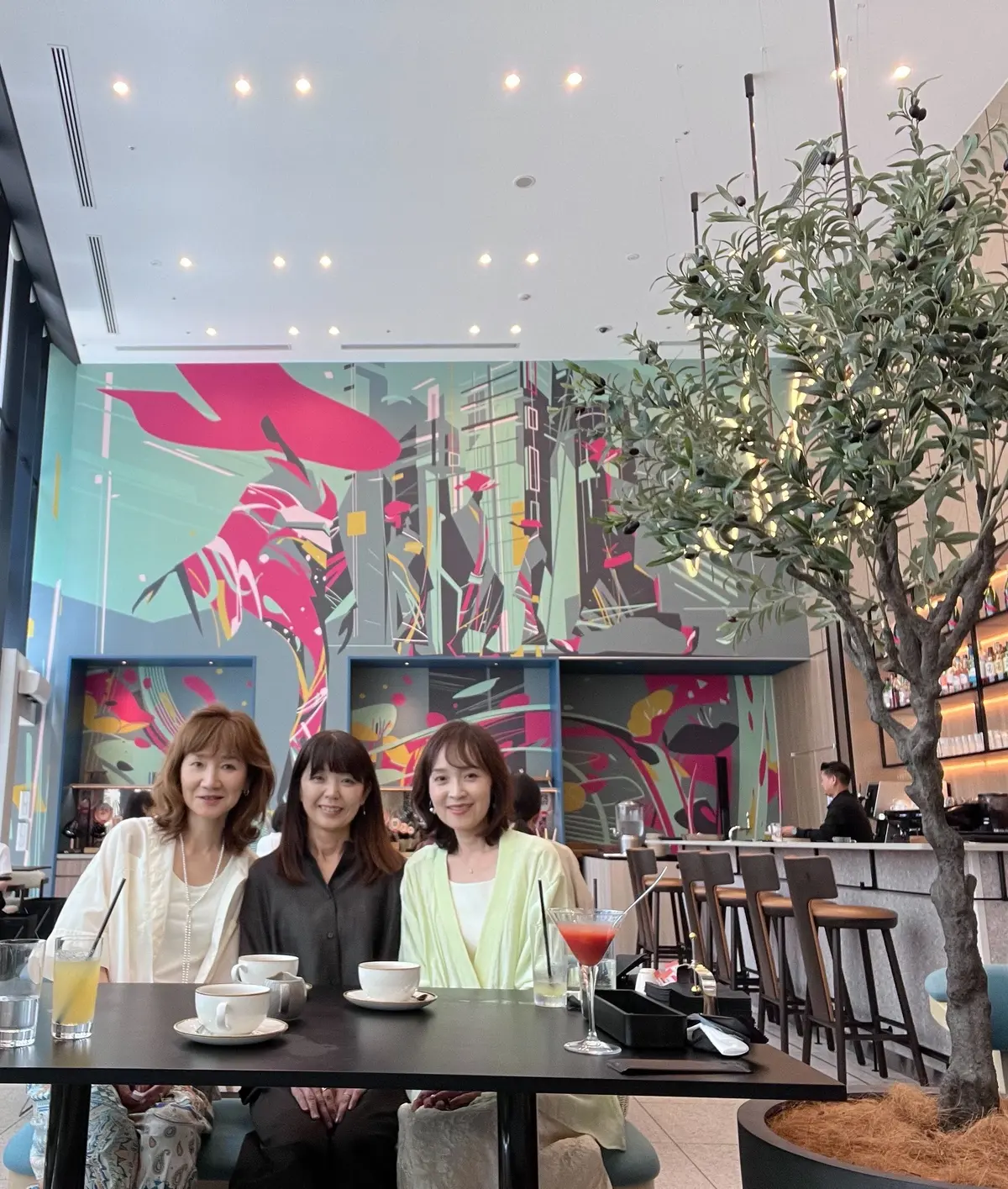 明るいカフェで、3人の女性がテーブルを挟んで座り、笑顔を見せている。背景にはカラフルな壁画と大きな観葉植物、窓から差し込む光が印象的。