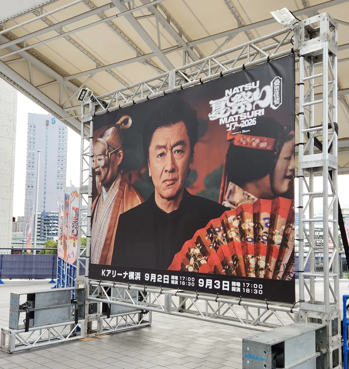 屋外ステージに設置された大型のイベントポスター。中央に男性の顔が大きく写り、背景には祭り風の装飾や人物が配置されている。「NATSU MATSURI 2026」の文字が目立つ。下部にはイベントの開催日時が記載されている。