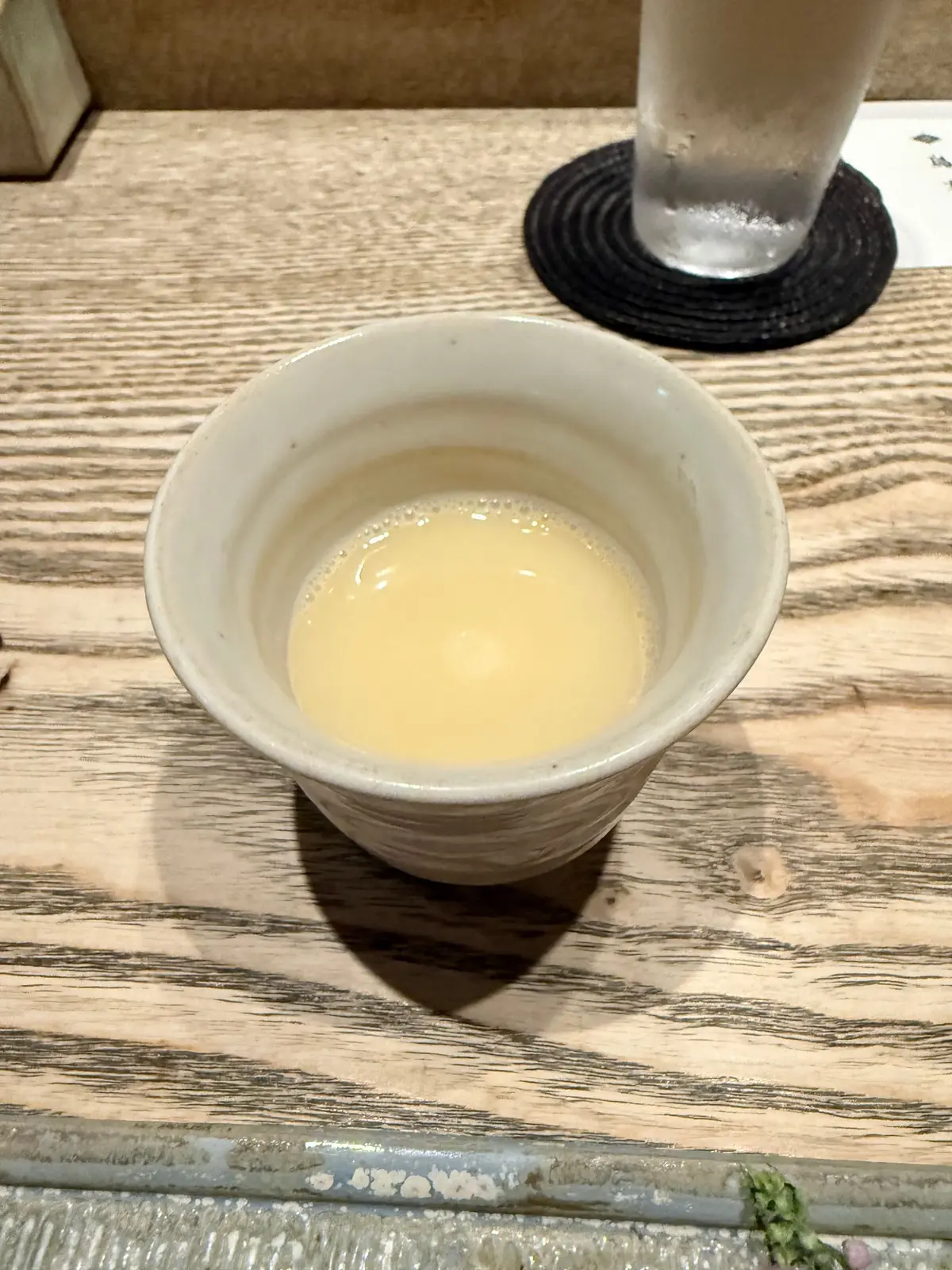 木製のテーブルに白い陶器の小鉢に入った淡い黄色いスープがあり、背景にガラスカップと黒いコースターが置かれている