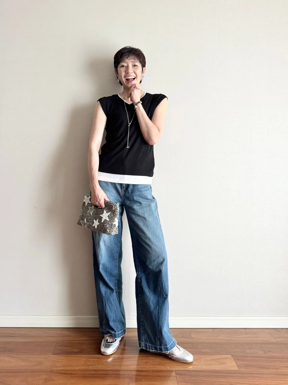 黒のノースリーブトップとデニムパンツを着た女性が、星模様のクラッチを左手に持って笑っている。木製の床の上に立っている。