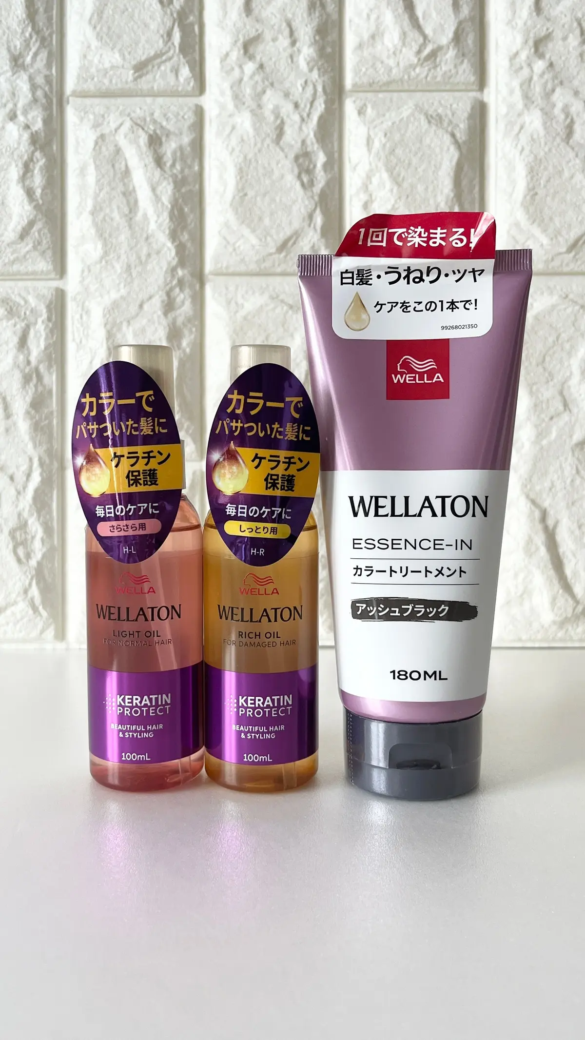 白いタイル壁に置かれた、ウェルアトン(Wellaton)のヘアケア製品3点。左から、赤いキャップのライトオイル、濃い紫色のリッチオイル、ピンク色のカラートリートメント。各製品のパッケージに日本語の文字表記あり。