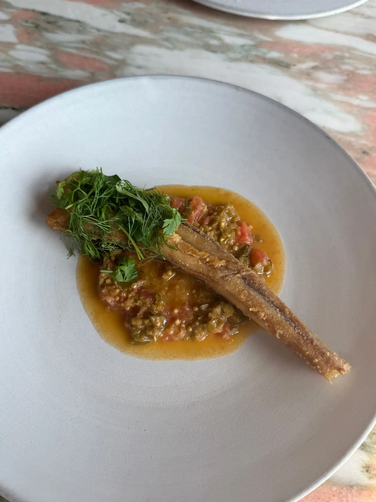 白磁の皿に盛り付けられた、揚げた魚の料理。魚は細長く、表面はカリカリ。周りにはトマトベースのソースと、緑色のハーブが添えられている。