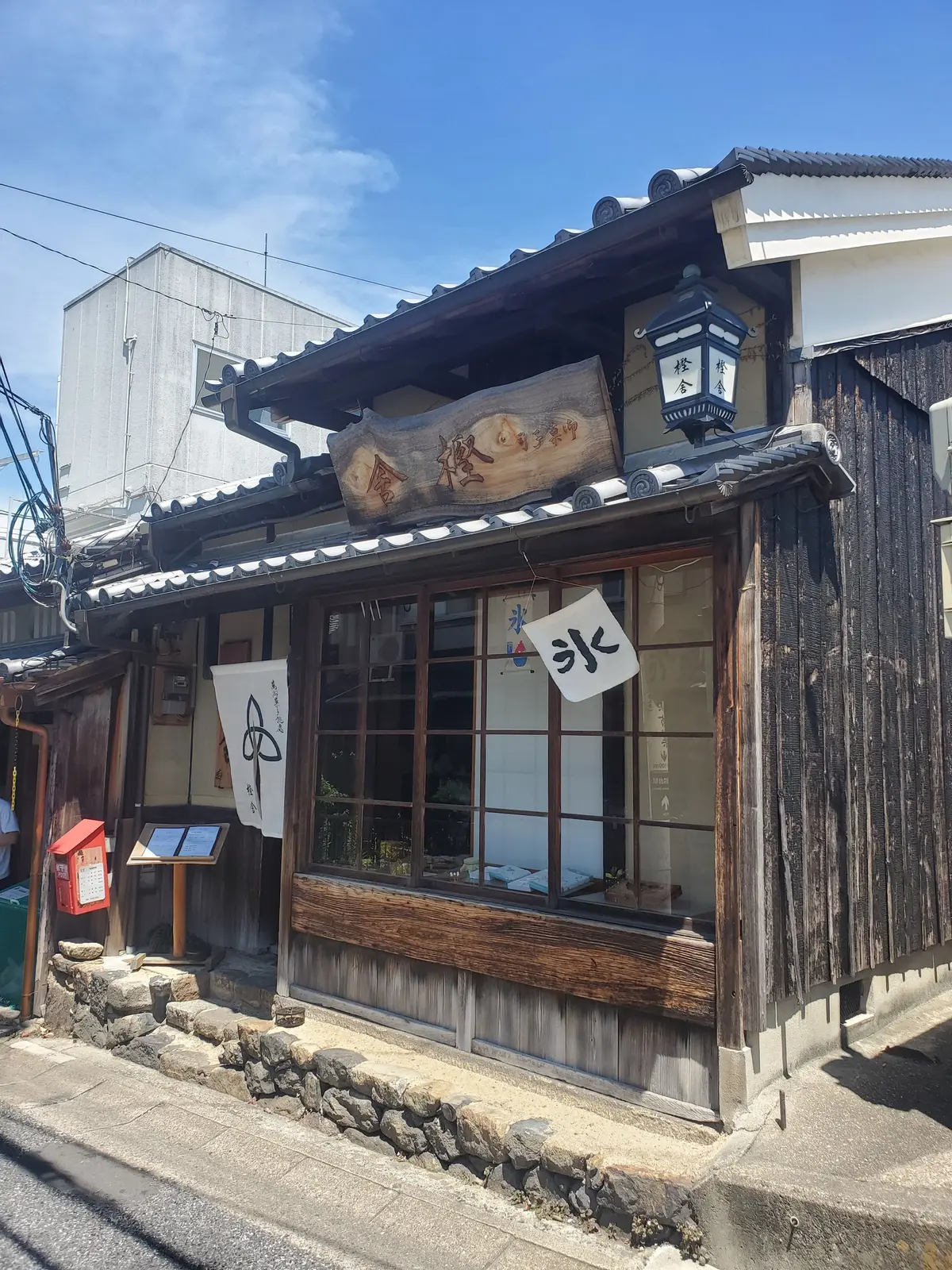木造の古い建物で、窓辺にはメニューらしきものが置かれている。看板は木の幹の形をしており、右には提灯のようなものがある。石畳の道と、周囲の建物が写っている。