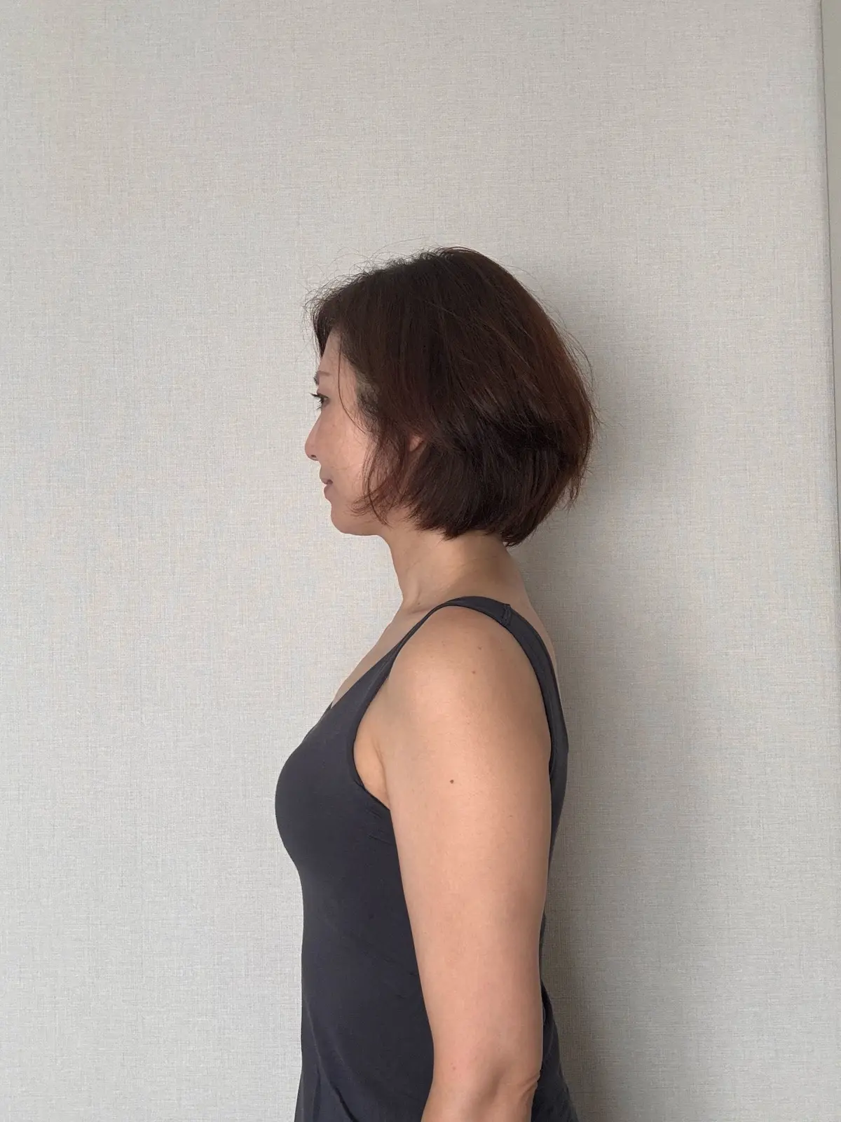 女性が横向きに立っている写真。茶色のボブヘアで、グレーのタンクトップを着用。背景はベージュの壁。