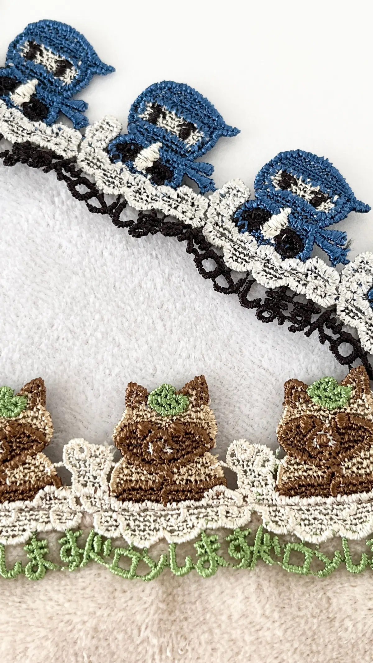 青と茶色の猫の刺繍が施されたレースのトリミングが2種類、白い背景に置かれている。上部は青い猫、下部は茶色の猫がそれぞれ配置されている。