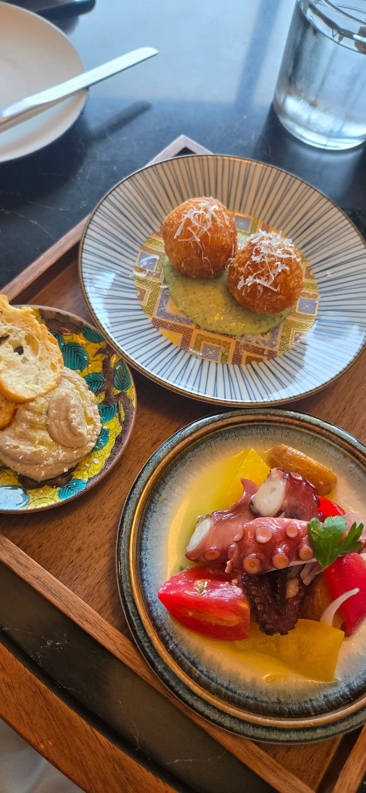 木製のトレイに盛り付けられた、3つの料理。上部は揚げたボール状の料理にチーズが乗せられ、下部左はホモウスにクラッカー、右はタコとトマト、オレンジのサラダ。それぞれ異なる模様の皿に盛り付けられている。
